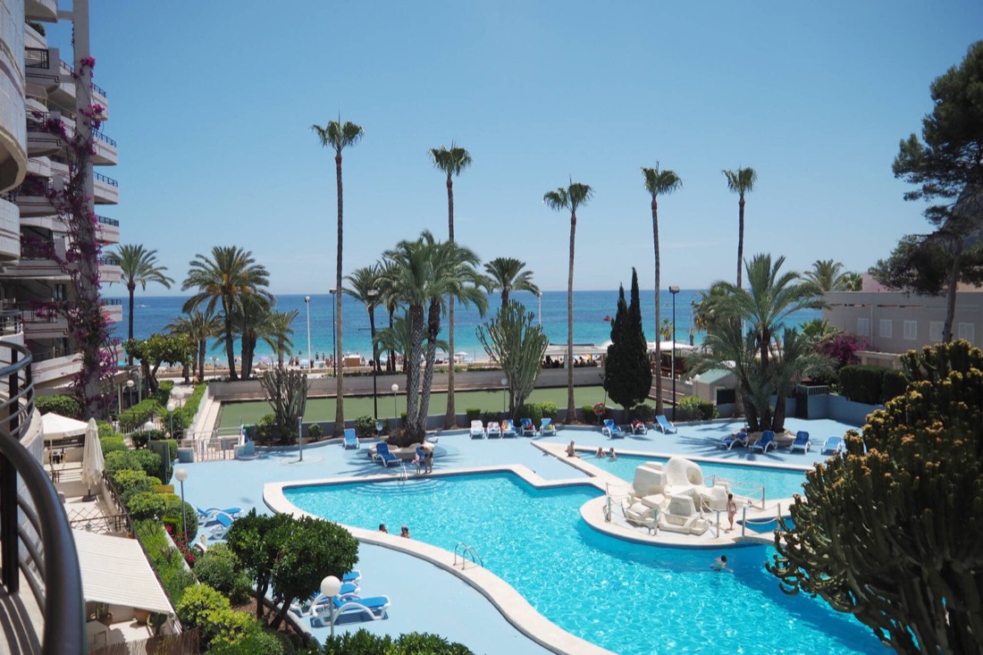Outdoor pool surrounded by palm trees, with Levante Beach beyond
