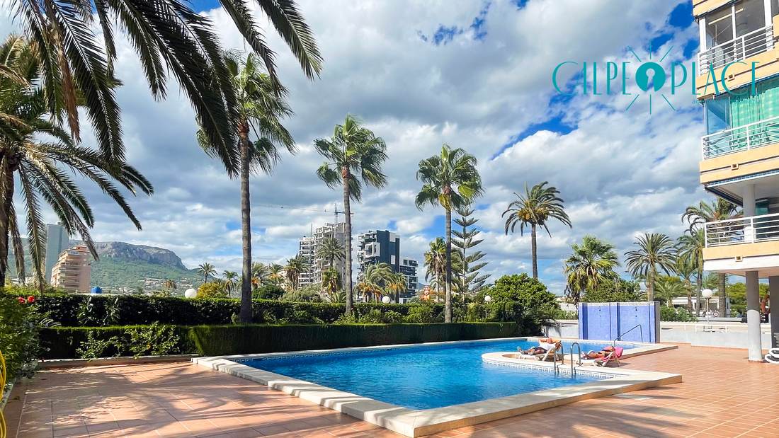 Piscina comunitaria de Oasis entre palmeras, con el Peñón de Ifach al fondo